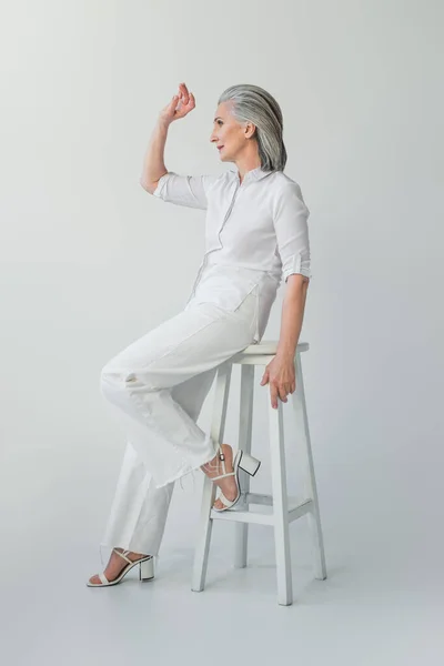 Зріла жінка в білому одязі дивиться на стілець на сірому фоні — стокове фото