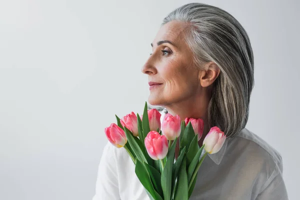 Mujer madura con tulipanes rosados mirando hacia otro lado aislados en gris - foto de stock