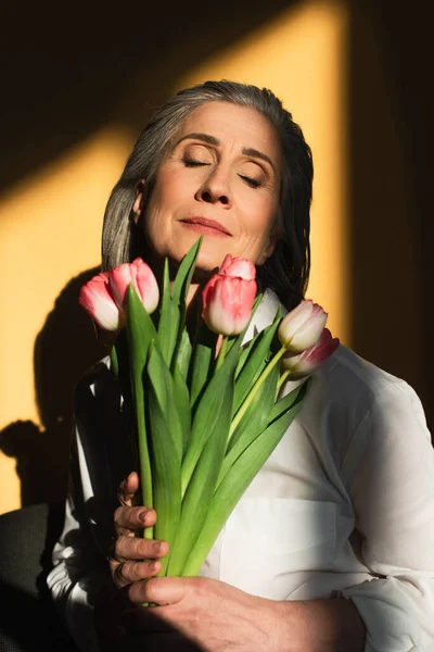 Ältere Frau mit geschlossenen Augen hält Tulpen auf gelbem Hintergrund mit Schatten — Stockfoto