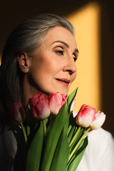 Middle aged woman holding pink tulips on yellow background with shadow — Stock Photo