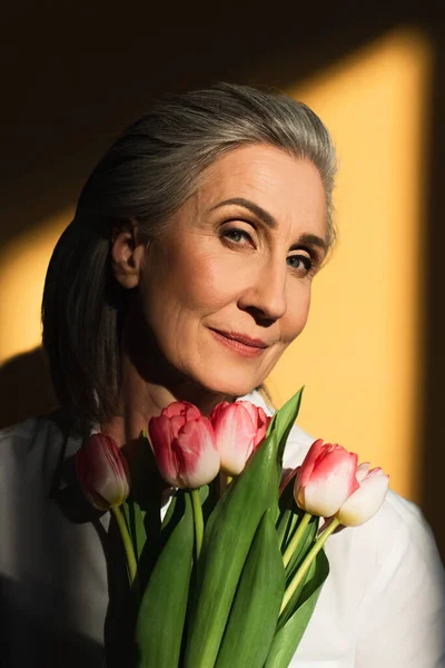 Ältere Frau hält Tulpen und schaut in die Kamera auf gelbem Hintergrund mit Schatten — Stockfoto