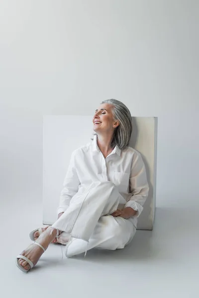 Mujer madura alegre con los ojos cerrados sentado cerca del cubo sobre fondo gris - foto de stock