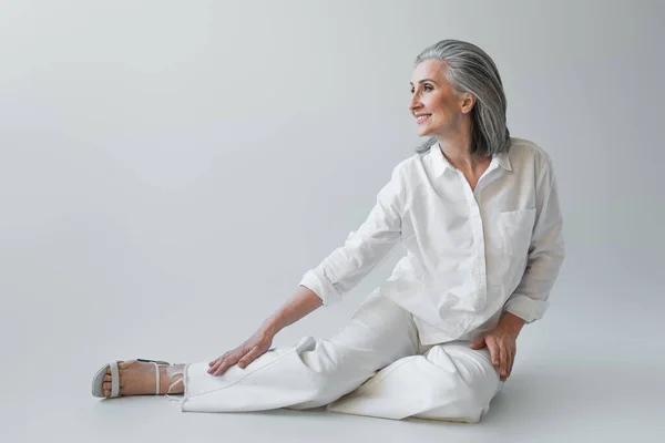 Allegro donna matura in abiti bianchi seduta su sfondo grigio — Foto stock