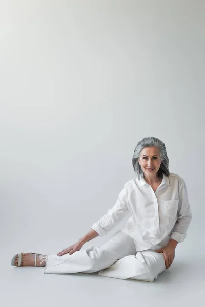 Femme aux cheveux gris souriant à la caméra tout en étant assis sur fond gris — Photo de stock