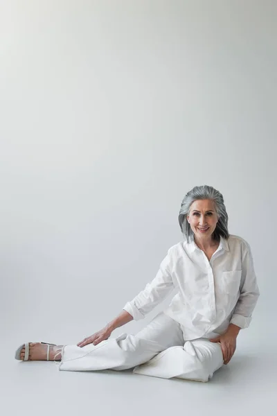 Mujer de pelo gris sonriendo a la cámara mientras está sentada sobre un fondo gris - foto de stock