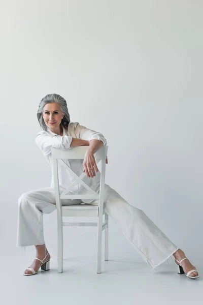 Frau in weißen Kleidern und Schuhen posiert auf Stuhl vor grauem Hintergrund — Stockfoto
