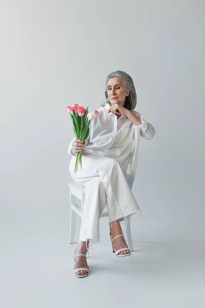 Ältere Frau schaut Blumen an, während sie auf einem Stuhl auf grauem Hintergrund sitzt — Stockfoto