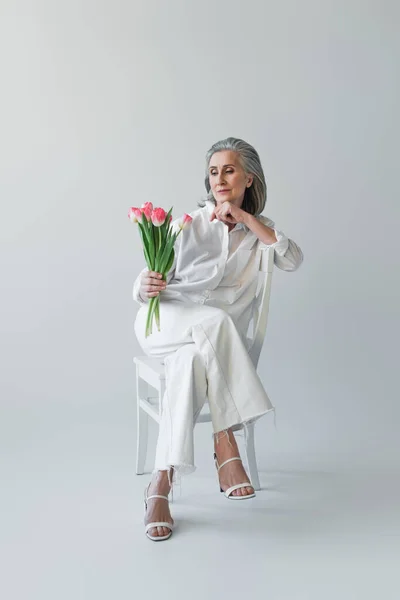 Mature woman looking at flowers while sitting on chair on grey background — Stock Photo