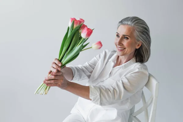 Mature woman smiling while looking at tulips on chair isolated on grey — Stock Photo