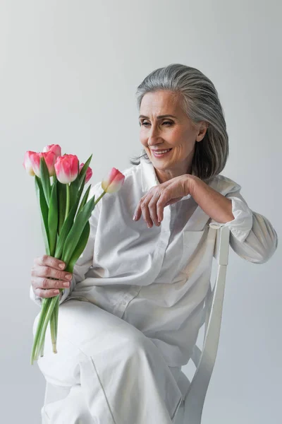 Donna dai capelli grigi in camicia bianca che guarda i fiori sulla sedia isolata su grigio — Foto stock