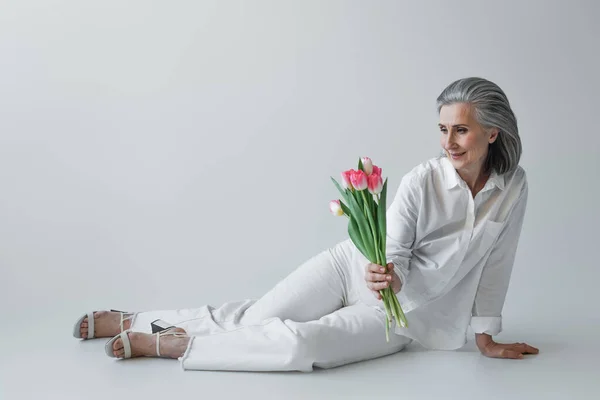 Улыбающаяся женщина в белой одежде держит тюльпаны на сером фоне — стоковое фото