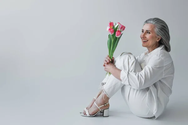 Fröhliche reife Frau hält einen Strauß Tulpen, während sie auf grauem Hintergrund sitzt — Stockfoto