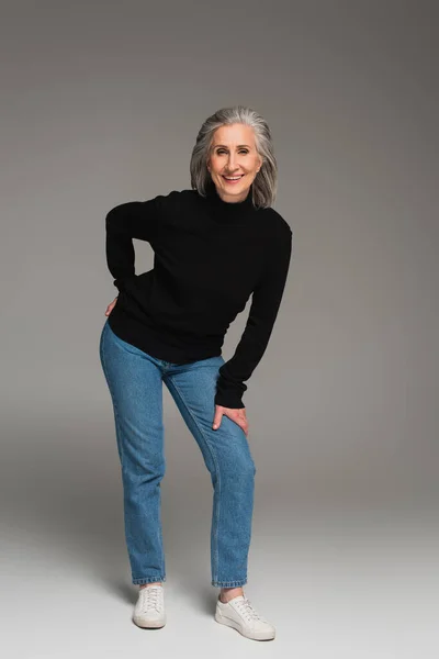 Полная длина улыбающейся женщины в свитере и джинсах на сером фоне — стоковое фото
