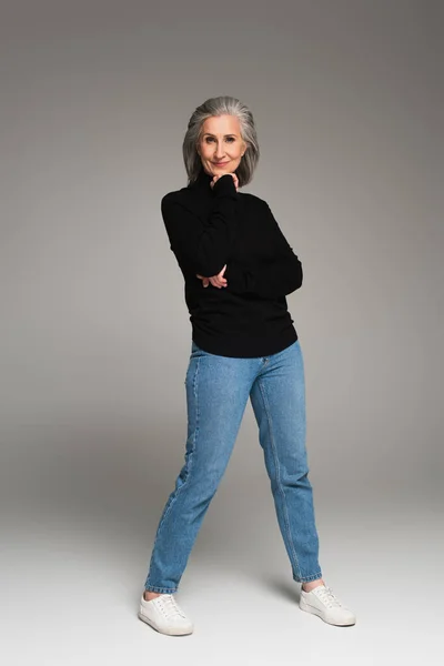 Comprimento total da mulher madura em jeans e gola alta olhando para a câmera em fundo cinza — Fotografia de Stock