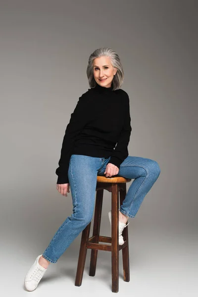 Femme mûre assise sur une chaise et souriant sur fond gris — Photo de stock