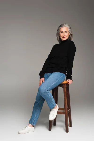 Mulher madura em jeans olhando para a câmera perto da cadeira no fundo cinza — Fotografia de Stock