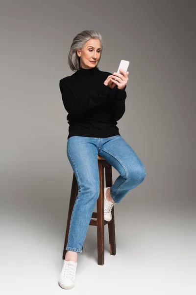 Mujer de mediana edad usando teléfono inteligente cerca de la silla sobre fondo gris - foto de stock