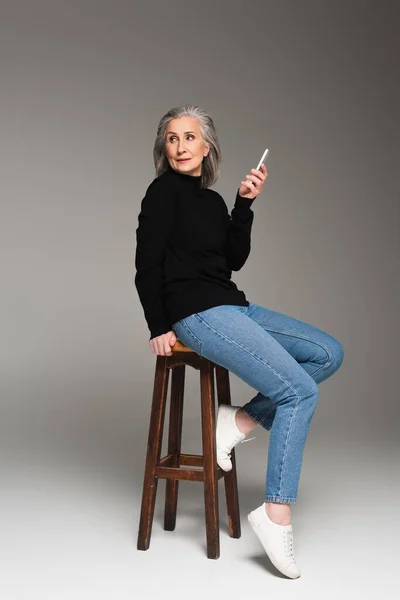 Mature woman in jeans holding smartphone on chair on grey background — Stock Photo