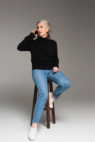 Grey haired woman talking on smartphone on chair on grey background — Stock Photo