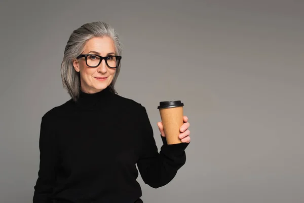 Donna matura con gli occhiali e maglione che tiene il caffè per andare isolato sul grigio — Foto stock