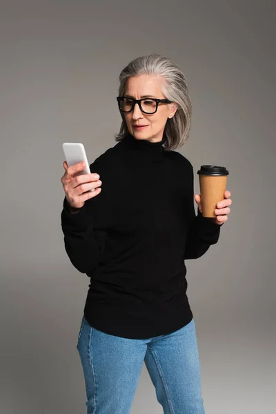 Reife Frau mit Brille hält Pappbecher in der Hand und benutzt Smartphone isoliert auf grau — Stockfoto