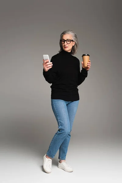 Mujer madura usando smartphone y sosteniendo la bebida para llevar sobre fondo gris - foto de stock