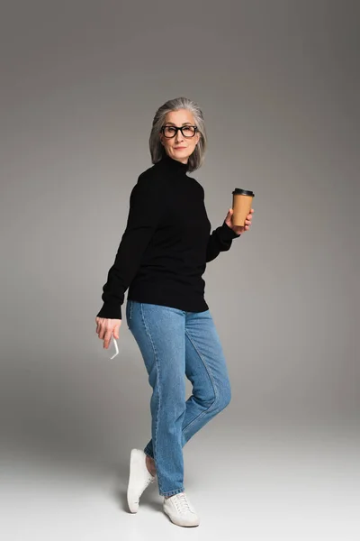 Femme aux cheveux gris dans des lunettes tenant du café à emporter et smartphone sur fond gris — Photo de stock