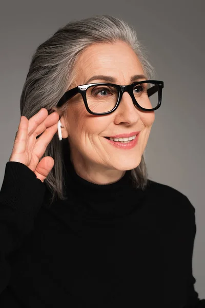 Femme mûre souriante dans des lunettes à l'aide d'un écouteur isolé sur gris — Photo de stock