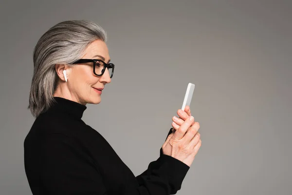 Зрелая женщина в наушнике с помощью смартфона, изолированного на сером — стоковое фото