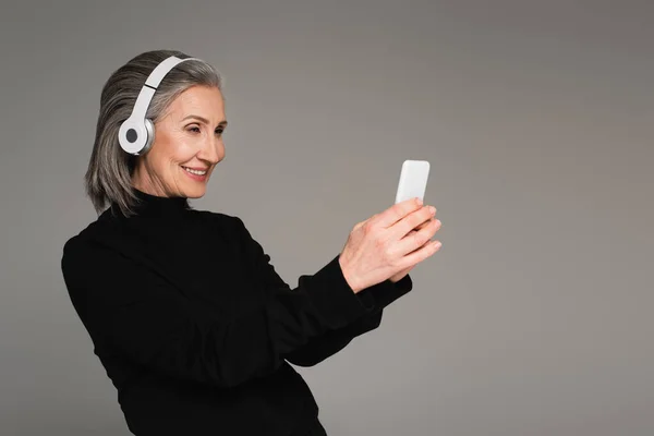 Sorridente donna matura in cuffia utilizzando smartphone isolato su grigio — Foto stock