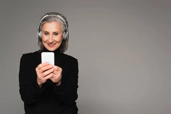 Middle aged woman in headphones using smartphone isolated on grey — Stock Photo