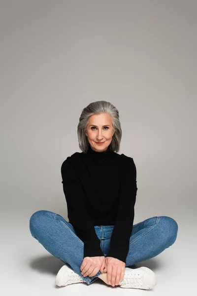 Mature woman with crossed legs sitting on grey background — Stock Photo