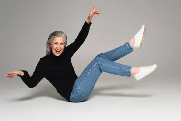 Позитивная зрелая женщина в джинсах сидит на сером фоне — стоковое фото