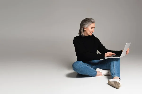 Mulher de gola alta e jeans usando laptop em fundo cinza — Fotografia de Stock