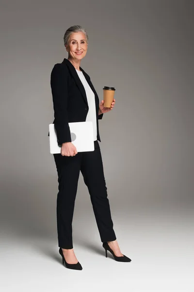 Empresária sorridente segurando bebida takeaway e laptop em fundo cinza — Fotografia de Stock