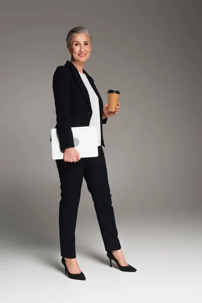 Smiling businesswoman holding takeaway drink and laptop on grey background — Stock Photo