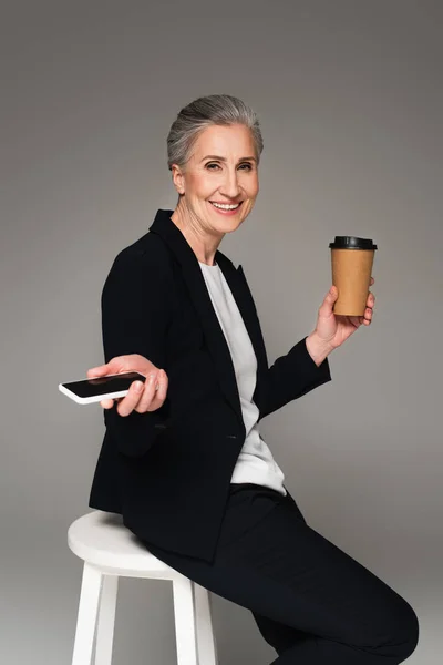 Femme d'affaires mature positive tenant smartphone et tasse en papier sur chaise sur fond gris — Photo de stock