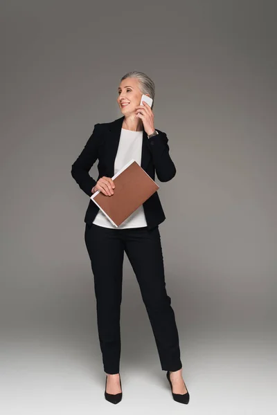 Усміхнена бізнес-леді з паперовою текою розмовляє на смартфоні на сірому фоні — стокове фото