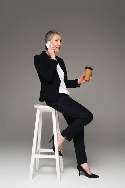 Donna d'affari sorridente con caffè da asporto che parla sul cellulare sulla sedia su sfondo grigio — Foto stock
