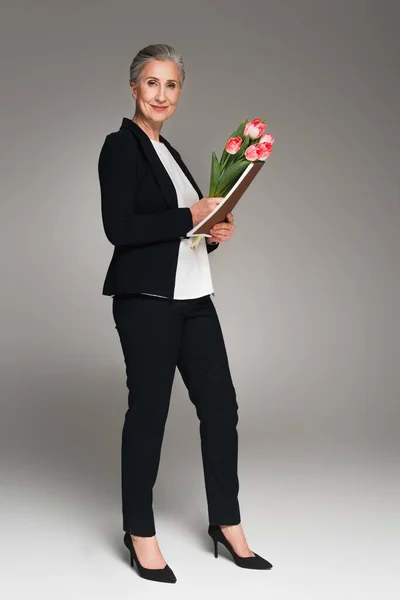 Smiling businesswoman holding paper folder and tulips on grey background — Stock Photo