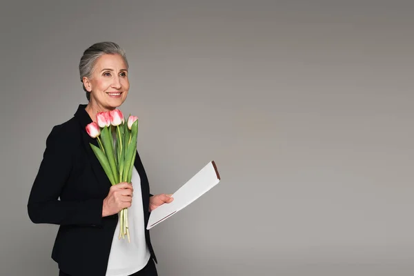 Улыбающаяся деловая женщина держит бумажную папку и тюльпаны изолированные на сером — стоковое фото