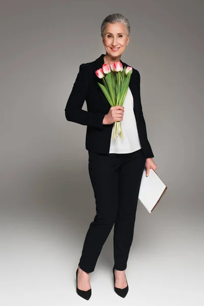 Positive businesswoman with paper folder holding tulips on grey background — Stock Photo