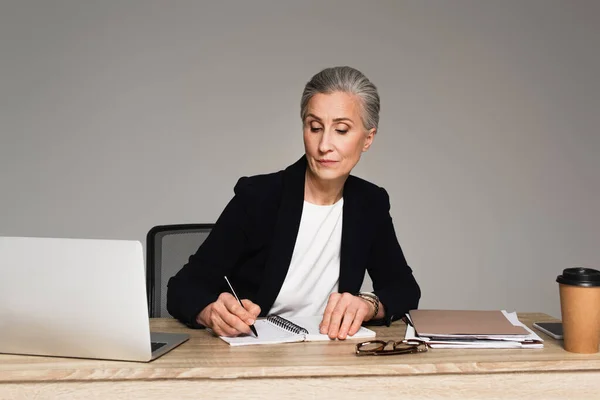 Businesswoman writing on notebook near gadgets and papers isolated on grey — Stock Photo