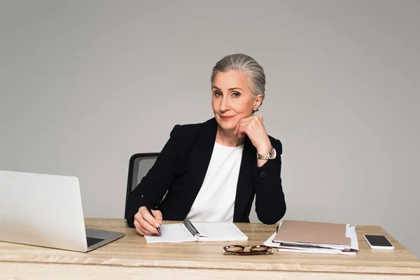 Madura empresaria escribiendo en cuaderno cerca de papeles y gadgets aislados en gris - foto de stock