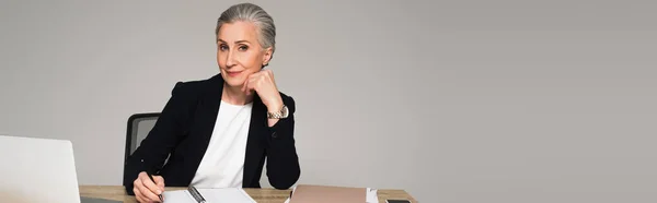 Mature manager writing on notebook near laptop and papers isolated on grey, banner — Stock Photo