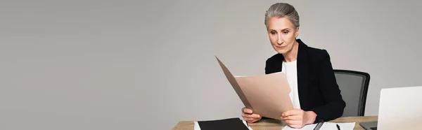 Femme d'affaires regardant dossier papier près d'un ordinateur portable sur la table isolé sur gris, bannière — Photo de stock