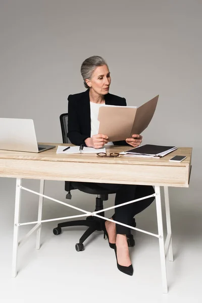 Reife Geschäftsfrau betrachtet Dokumente in der Nähe von Gadgets und Brillen auf einem Tisch auf grauem Hintergrund — Stockfoto