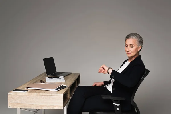Donna d'affari che guarda orologio da polso vicino a dispositivi sul tavolo su sfondo grigio — Foto stock