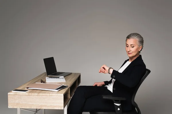 Mujer de negocios mirando el reloj de pulsera cerca de los dispositivos en la mesa sobre fondo gris - foto de stock