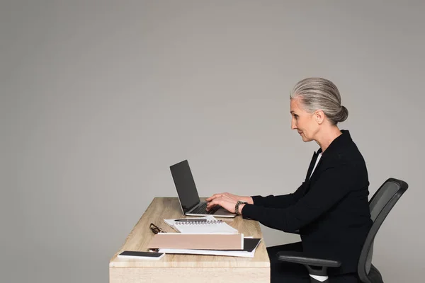 Вид сбоку на предпринимательницу, использующую ноутбук рядом с бумагами, изолированными на сером фоне — стоковое фото
